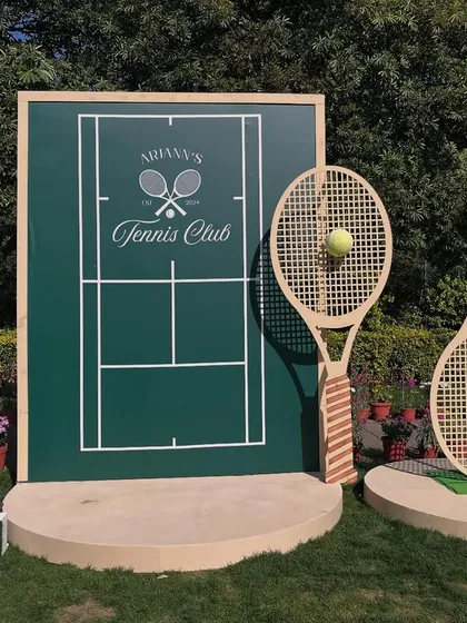 A sophisticated Tennis Club theme for a first birthday. The main photo backdrop features a custom tennis court design, oversized rackets, and a grass-covered number '1'.