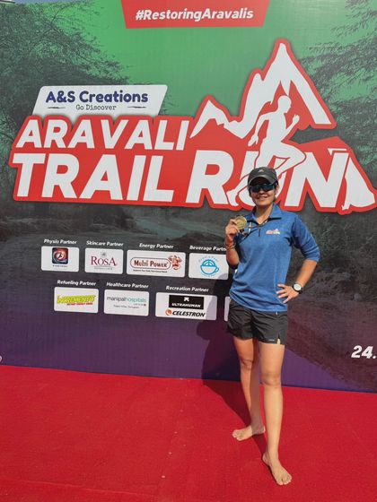 A finisher proudly posing with her medal in front of the Aravali Trail Run event backdrop.