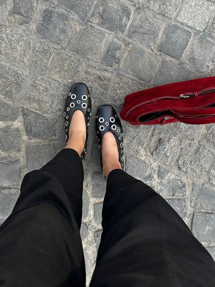 A street-style view of the black eyelet ballet flats paired with a red suede bag. This is how I love to see my pieces worn, as part of a real-life look that is both stylish and practical.
