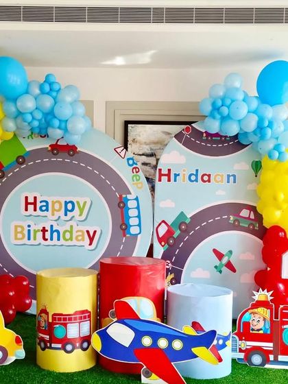 A transportation-themed birthday party perfect for a little one who loves things that go. The setup includes cars, planes, and trucks against a roadway backdrop with bright, primary-colored balloons.