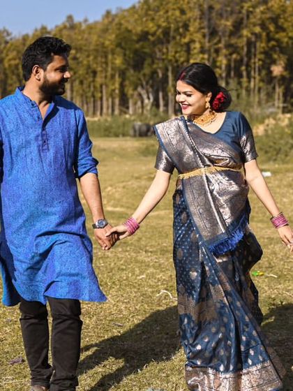 A full-length shot of the couple in their traditional outfits, walking through an open field. This showcases the elegance of the saree against a rustic, natural landscape.