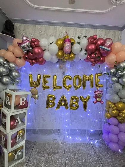 Even a small drawing room can be decorated beautifully to welcome your new baby. This setup includes a balloon arch, 'Welcome Baby' foil letters, and a backdrop with fairy lights for a warm and cozy celebration at home.