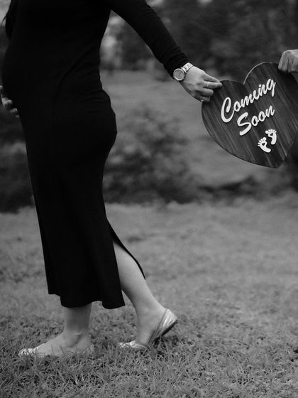 A black and white detail shot of the mom-to-be walking while holding a "Coming Soon" sign. The motion and composition create a feeling of moving forward into a new chapter.