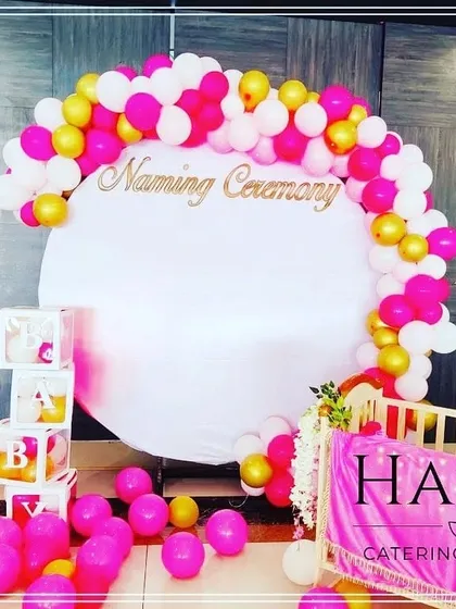 A bright and cheerful naming ceremony setup. This design uses a vibrant pink, white, and gold balloon arch against a circular backdrop, complemented by "BABY" blocks and a decorated cradle.