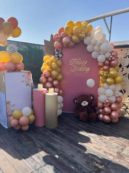 A beautiful terrace birthday decoration with a warm, earthy color palette. The setup includes a pink backdrop, a balloon arch in gold, white, and rose gold, and a cute teddy bear prop.