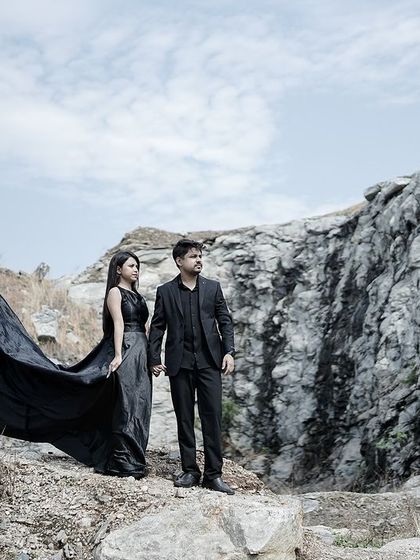 A dramatic pre-wedding portrait in a rocky landscape, with the couple in black formal wear and the wind catching the long trail of her gown.