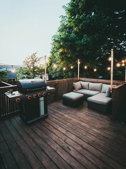 This classic wooden deck is perfect for entertaining, featuring a comfortable L-shaped sofa and a barbecue grill. The string lights add a warm, festive glow for evening get-togethers.