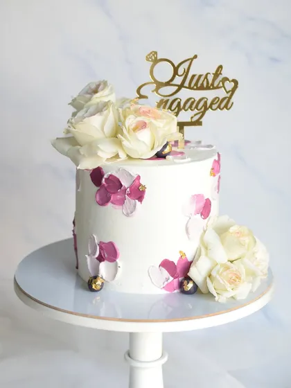 A simple yet beautiful single-tier engagement cake. The design features a palette-knife texture in shades of purple and pink, topped with fresh flowers and a "Just Engaged" topper.