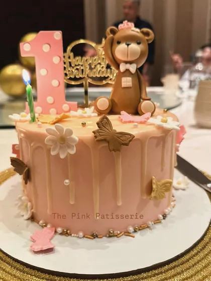 A stylish drip cake for a first birthday. This pink cake features a white chocolate drip, a fondant teddy bear, and delicate butterflies and flowers.