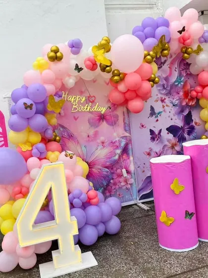 A close-up of the butterfly theme backdrop, featuring large butterfly wing cutouts, a marquee number '4', and a balloon arch in pink, purple, and yellow.