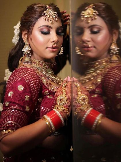 A stunning bridal portrait reflecting in the mirror. The makeup features a warm, bronzed smokey eye and a glossy nude lip, paired with a traditional matha patti and gajra braid for a complete goddess look.