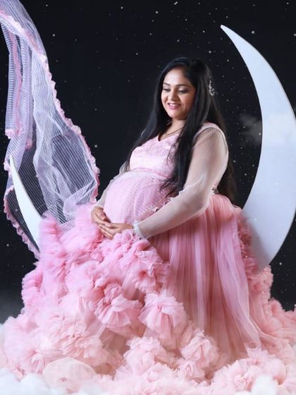 A dreamy, celestial-themed maternity shoot. The mom-to-be is seated on a crescent moon prop, wearing a pink ruffled gown that looks like a cloud.