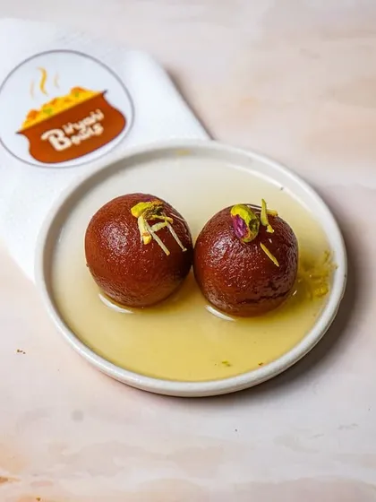 A dark and moody shot of two gulab jamuns in syrup, with the brand's napkin subtly placed. This lighting style adds a touch of elegance and drama to a traditional dessert.