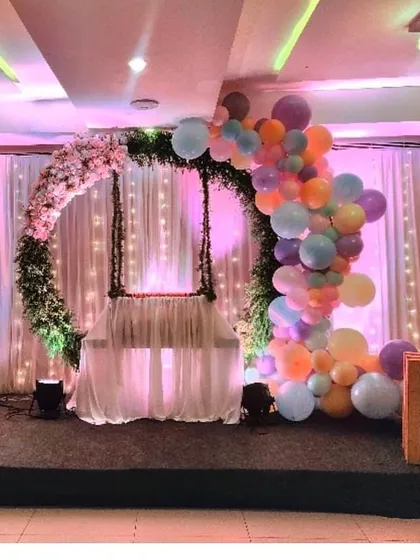 An elegant and serene cradle ceremony decoration. The setup features a beautifully decorated swing at the center of a floral ring, with soft curtain backdrops and a cascade of pastel balloons, creating a blessed ambiance.