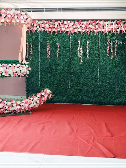 A naming ceremony stage with a floral crescent moon cradle set against a lush green wall. The backdrop is adorned with hanging pink floral strings for a delicate touch.