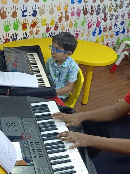 An older student plays the piano while a younger one watches with curiosity. Our classes often inspire the next generation of musicians.