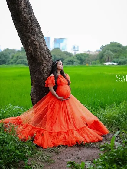 Stand out in a field of green with this breathtaking orange ball gown. The voluminous skirt and rich color create a beautiful contrast with nature, perfect for a serene and powerful outdoor maternity portrait.