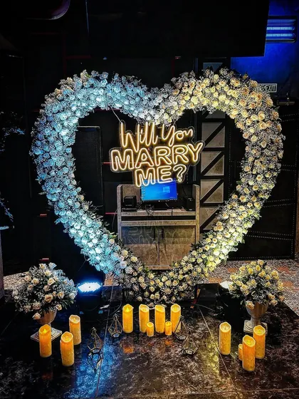 An elegant and dreamy proposal setup featuring a heart-shaped arch of pure white flowers. The glowing candles and central neon sign create a sophisticated and magical ambiance in a darker, intimate setting.