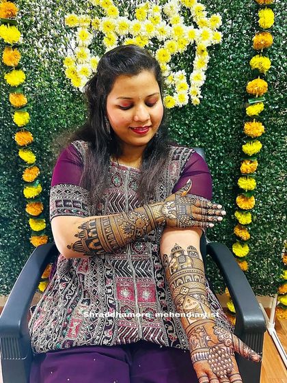 A happy bride seated in front of a festive backdrop, proudly displaying her full bridal mehendi.