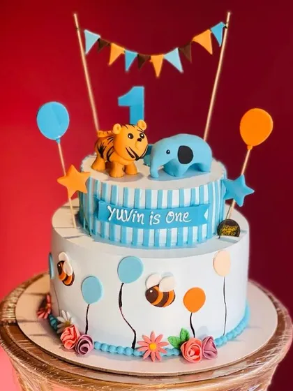 A sweet two-tier cake for a first birthday celebration. This design includes cute animal toppers like a tiger and elephant, with balloons and a festive banner.