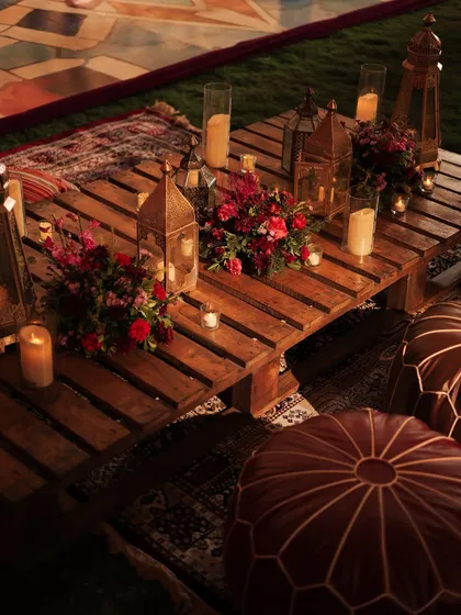 An aerial view of the rustic picnic-style table, showing the arrangement of lanterns, candles, and floral bunches. The layered rugs and leather ottomans add texture and comfort.