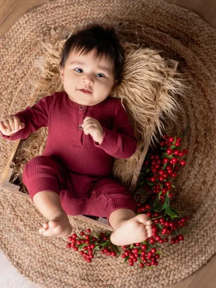 A happy baby in a maroon romper lies in a wooden crate on a jute rug. The simple, rustic props and pops of red berries create a warm and earthy portrait.