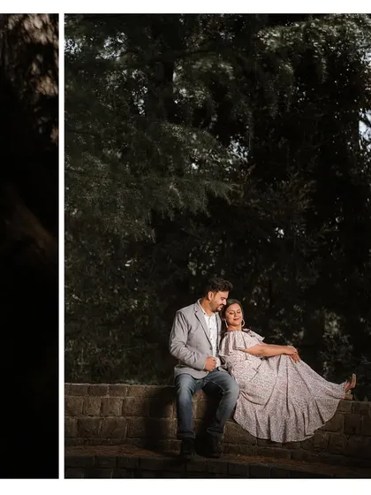A couple sits on a stone wall in a forest, sharing a quiet moment. The play of light and shadow adds a moody, cinematic quality to the portrait.