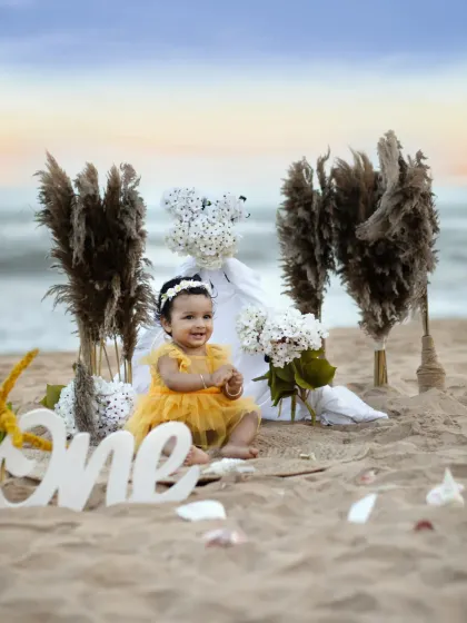 A happy baby girl claps her hands with excitement during her "One" birthday photoshoot on the sand, surrounded by boho-style props.