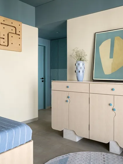 This son's bedroom in the 'Soft Serve' project uses soft blue tones and light wood to create a calm and playful atmosphere. The custom desk and overhead storage unit feature circular cutouts, adding a touch of modern geometry.