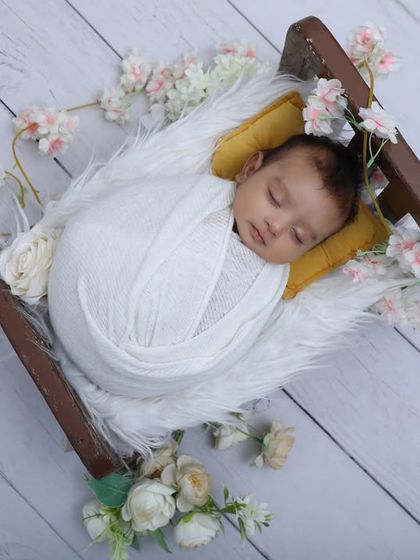 Dreaming peacefully. This overhead shot shows a newborn swaddled in white, resting in a miniature sleigh on a white wood-paneled floor, surrounded by delicate pink and white flowers.