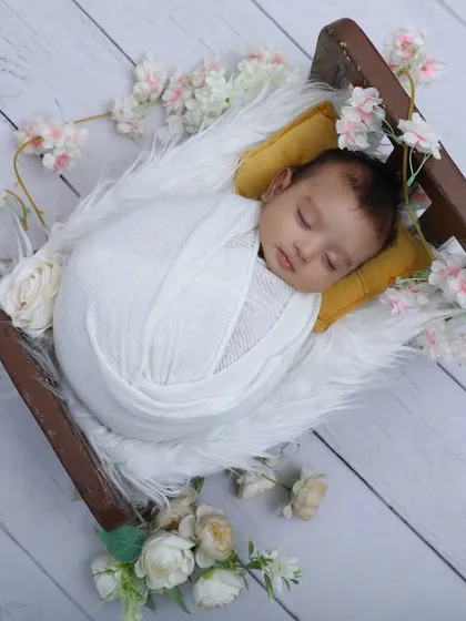 Dreaming peacefully. This overhead shot shows a newborn swaddled in white, resting in a miniature sleigh on a white wood-paneled floor, surrounded by delicate pink and white flowers.
