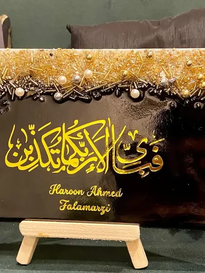 This personalized plaque features the verse "Fabi ayyi ala i rabbikuma tukazziban" in bold yellow calligraphy on a black background. The top is adorned with a textured mix of gold and silver embellishments, and it includes the client's name for a personal touch.