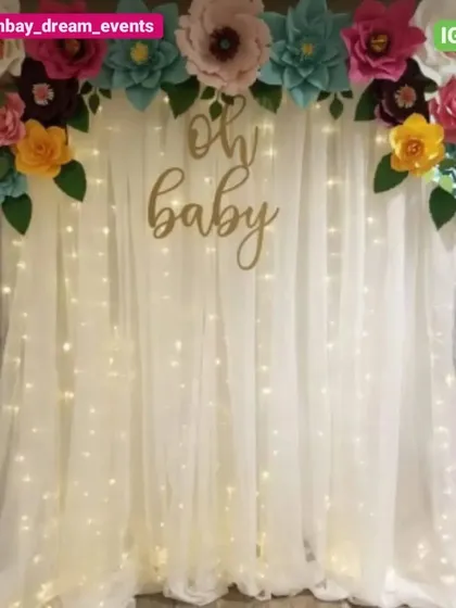 A beautiful 'Oh Baby' backdrop featuring a white curtain with fairy lights. The top is adorned with a garland of colorful, handcrafted paper flowers, adding a unique and artistic touch.