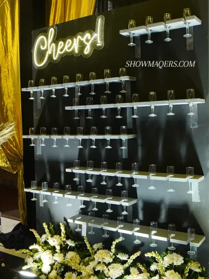 A closer look at the champagne wall, showcasing its simple yet effective design. The neon "Cheers!" sign adds a fun and celebratory touch to the black and gold theme.
