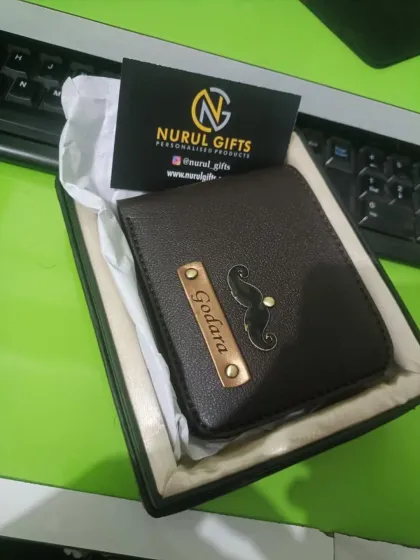 A close-up of a dark brown leather wallet, personalized with the name 'Godara' and a mustache charm, presented in its gift box.