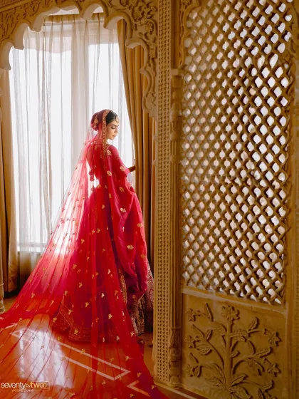 A bride in her stunning red wedding lehenga, with a long, dramatic veil, gazes out of a window framed by intricate jali work. A portrait of classic bridal beauty.
