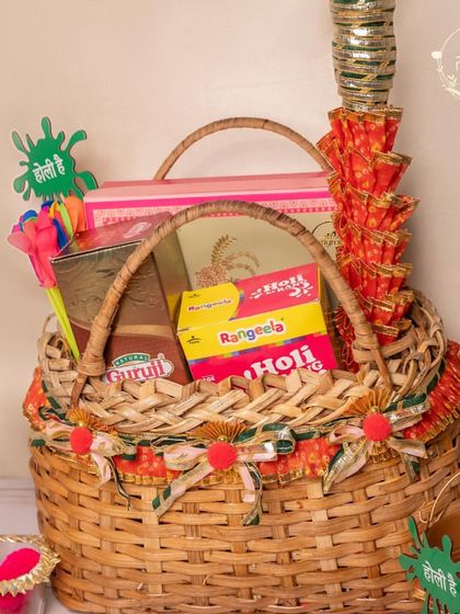 A classic cane basket Holi hamper, filled with all the essentials like organic colours, pichkaris, and boxes of sweets and snacks for a complete celebration.