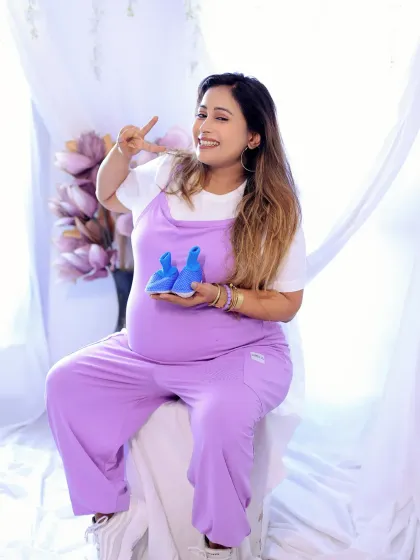 A fun and playful studio photo. The mother-to-be is dressed in cute lavender overalls and holds up a pair of tiny blue baby shoes.