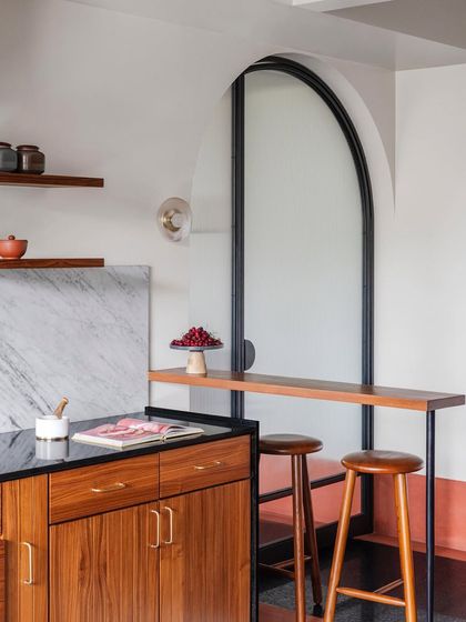 This kitchen view shows a custom bar counter with stools, set against an arched doorway. The mix of timber, marble, brass, and metal creates a layered and sophisticated space.