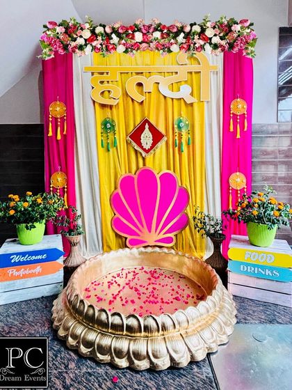 A compact and colorful Haldi setup for a home function. I used a 'Haldi' sign in Hindi, a seashell cutout, and quirky painted boxes to create a fun and vibrant photo corner.