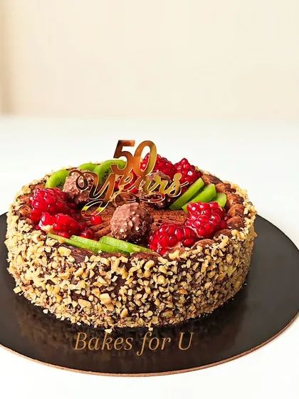 A whole wheat cake created for a 50th anniversary celebration. The sides are coated in chopped nuts, and the top is decorated with fresh raspberries, kiwi, and Ferrero Rocher chocolates.