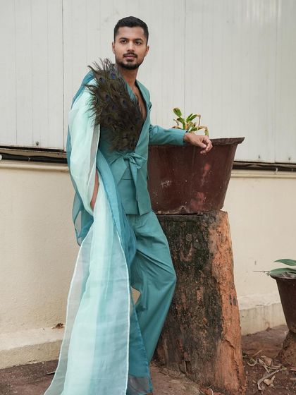 An avant-garde look featuring a teal suit with a flowing sash and peacock feathers. This is for high-concept, editorial fashion.