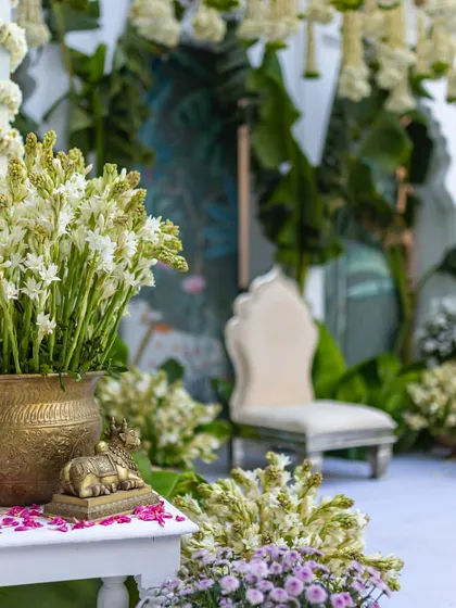 A close-up of the details in a traditional decor setup. A brass vessel holds fresh tuberose, placed next to a small Nandi statue, with scattered flower petals adding color. These elements create a sense of serenity and devotion.