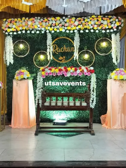 An elegant naming ceremony stage with a greenery backdrop and a beautifully decorated wooden cradle. The floral arrangements on stands add a touch of class.