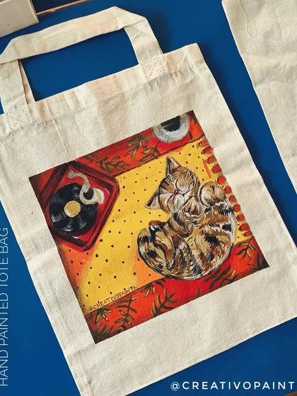 For all the cat lovers out there. This is a hand-painted tote bag featuring a cozy cat napping next to a record player, perfect for carrying your things in style.