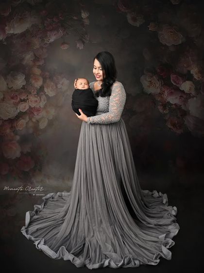 An elegant mother and newborn portrait. Dressed in a beautiful grey gown, she holds her baby against a dark, floral backdrop, creating a timeless and artistic image.
