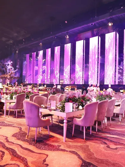 A beautifully arranged dining space with plush velvet chairs and mirrored tables, reflecting the dramatic purple lighting and creating an atmosphere of pure luxury.