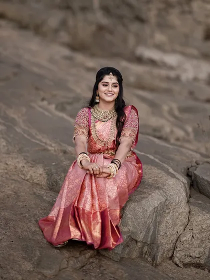 A beautiful bridal portrait of the bride seated on a rock. Her joyful smile and stunning pink saree stand out against the natural, rugged background.