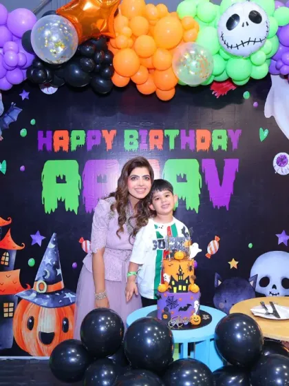 A fantastic Halloween-themed birthday setup. The backdrop features a haunted house and spooky characters, all framed by a balloon arch in classic Halloween colors.