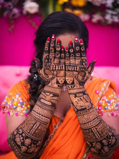 A full view of the bride's hands, held up to her face. The bright red nails provide a stunning contrast to the dark henna.